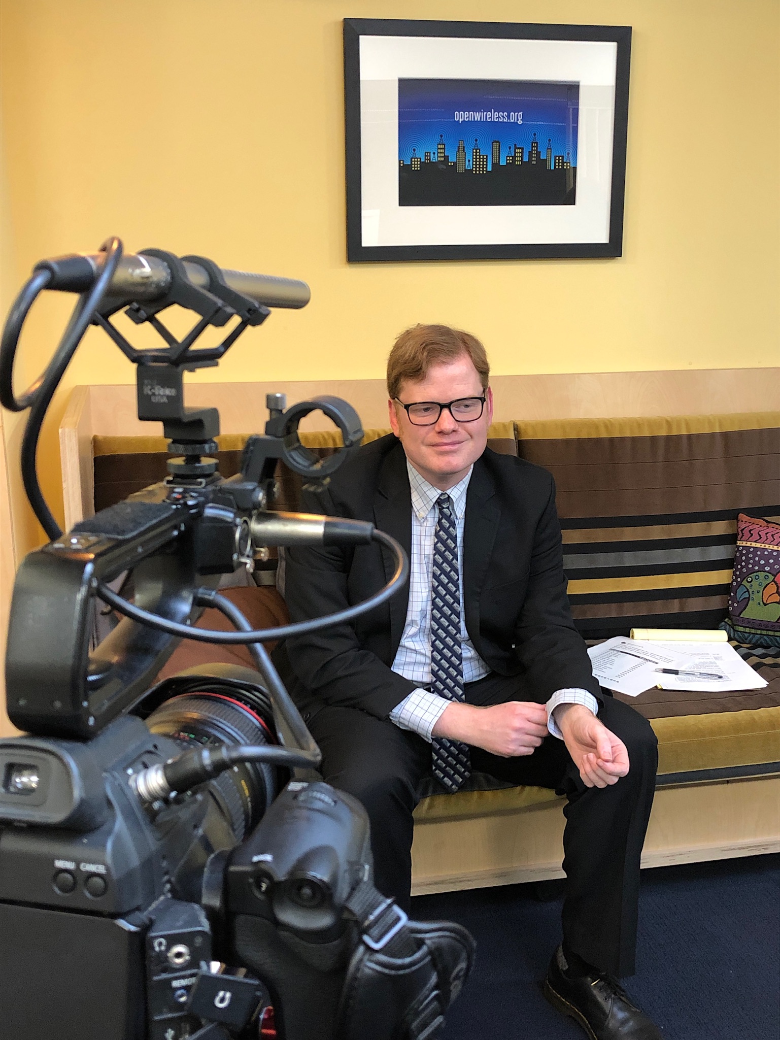 Elliot is wearing his glasses, a suit and a blue tie underneath an illustration for Open Wireless.