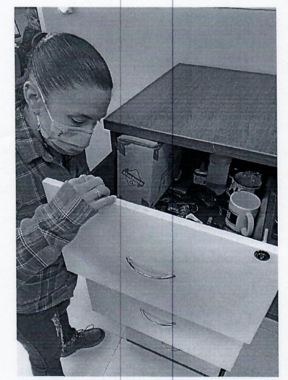 A badly photocopied image of a person wearing a COVID mask searching a filing cabinet. The top drawer has coffee mnugs and other related items. 