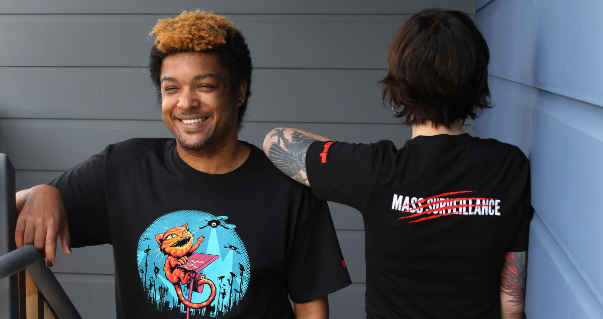 Get a Claw Back t-shirt at the Copper member level and above! Two people wear EFF Claw Back member t-shirts. The front shows a cat swatting at spy cameras and the back says “Mass Surveillance” with red claw marks through it.