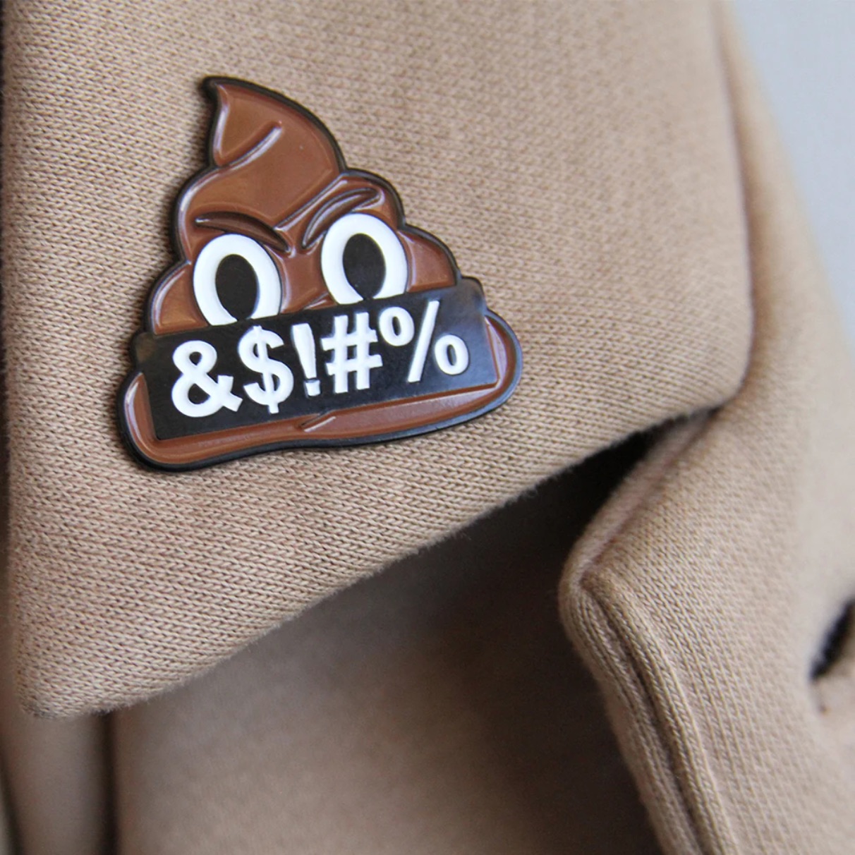 A close-up of an enamel pin on the lapel of a tan jacket. The pin depicts an angry poop emoji holding a black banner reading "&$!#%". A close-up of an enamel pin on the lapel of a tan jacket. The pin depicts an angry poop emoji holding a black banner reading "&$!#%".